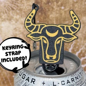 May include: A black and gold bull-shaped keychain charm with a keyring strap. The charm is placed on top of a silver can. The text "KEYRING STRAP INCLUDED!" is in a white circle on the left side of the image.