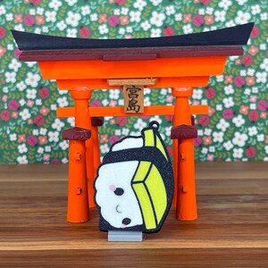 Pu&ograve; includere: Un piccolo ciondolo sushi stampato in 3D, in stile cartone animato, con un design giallo e nero. Il ciondolo &egrave; appeso a un torii, un tradizionale portale giapponese rosso e arancione.