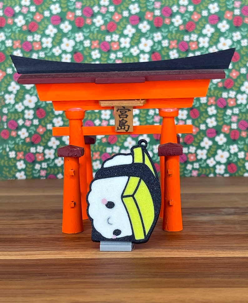 Pu&ograve; includere: Un piccolo ciondolo sushi stampato in 3D, in stile cartone animato, con un design giallo e nero. Il ciondolo &egrave; appeso a un torii, un tradizionale portale giapponese rosso e arancione.