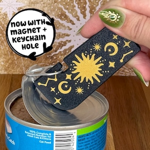 Pode incluir: Um abridor de latas preto para comida de gato com um design dourado de sol, lua e estrelas. O texto "now WITH magnet + keychain HOLE" está em um círculo branco. O abridor de latas está sobre uma lata de comida de gato.