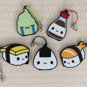 Pu&ograve; includere: Cinque portachiavi colorati con personaggi di cibo giapponese. I portachiavi sono fatti di feltro e includono un rotolo di sushi, una palla di riso, una bottiglia di salsa di soia, un pezzo di alga e un pezzo di sushi con un gambero in cima.