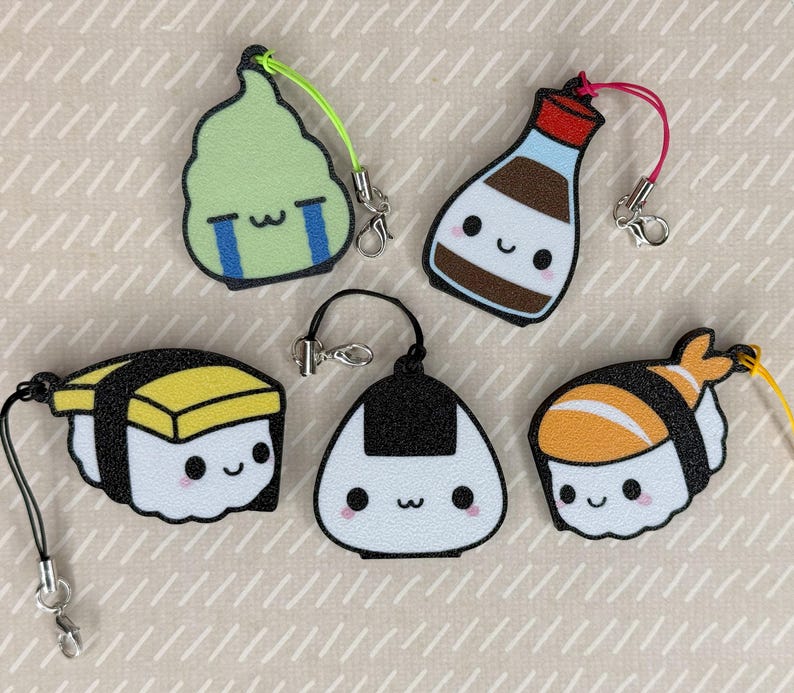 Pu&ograve; includere: Cinque portachiavi colorati con personaggi di cibo giapponese. I portachiavi sono fatti di feltro e includono un rotolo di sushi, una palla di riso, una bottiglia di salsa di soia, un pezzo di alga e un pezzo di sushi con un gambero in cima.
