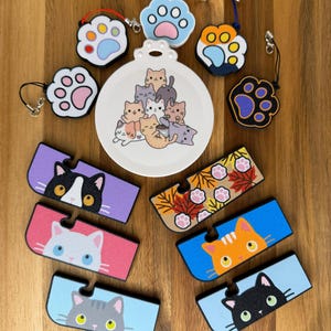 May include: A collection of cat-themed accessories including a round white ornament with a pile of cartoon cats, five paw-shaped charms with various color schemes, and six rectangular phone stands featuring cat faces and autumn leaves.
