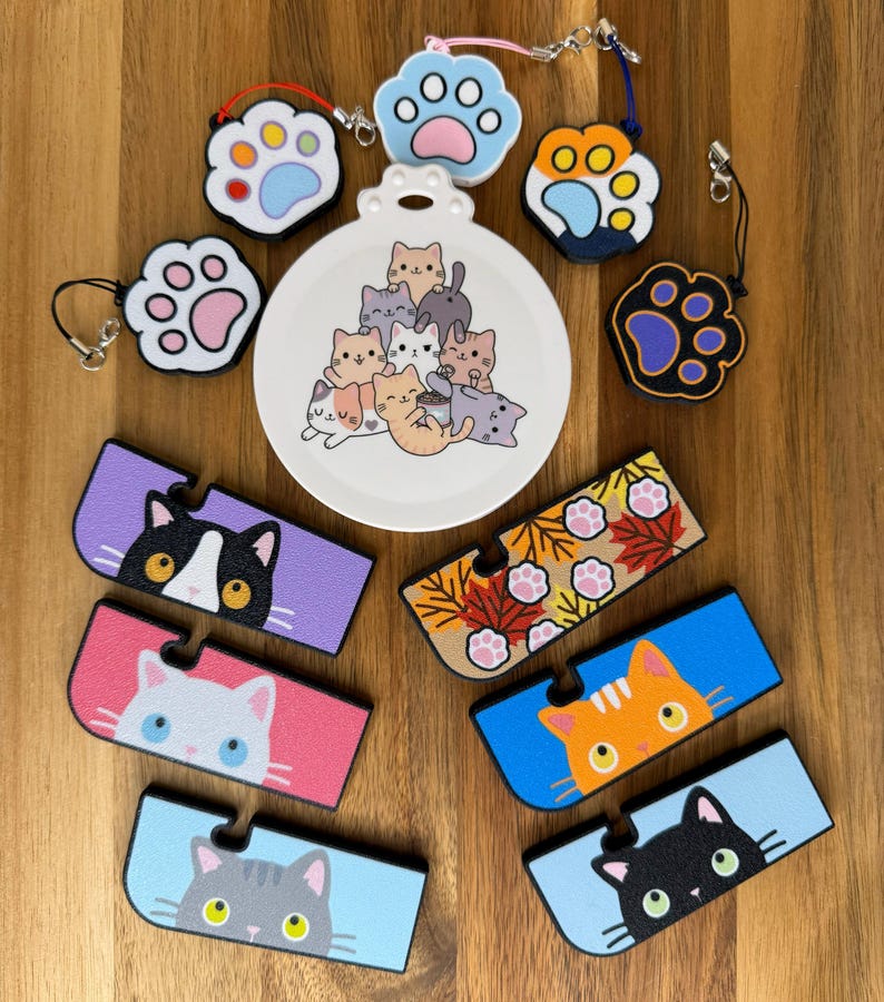 May include: A collection of cat-themed accessories including a round white ornament with a pile of cartoon cats, five paw-shaped charms with various color schemes, and six rectangular phone stands featuring cat faces and autumn leaves.