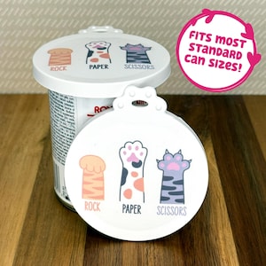 May include: Two white circular silicone can lids featuring a graphic of three cat paws labelled Rock, Paper, and Scissors. One lid is placed on a metal can, while the other is positioned in front. A pink circular graphic states FITS MOST STANDARD CAN SIZES!