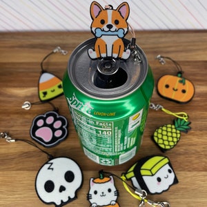 Pu&ograve; includere: Una collezione di nove ciondoli a tema Halloween, tra cui una zucca, un teschio, un gatto, un'impronta di zampa, un ananas, un rotolo di sushi, un mais dolce e un corgi. I ciondoli sono appesi a una lattina di Sprite.