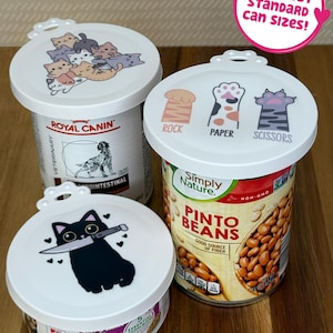 May include: Three white circular silicone can lids with printed designs. One lid features a pile of colorful kittens, another shows cat paws labeled Rock, Paper, Scissors, and the third displays a black cat holding a knife. A pink sticker reads FITS MOST STANDARD CAN SIZES!