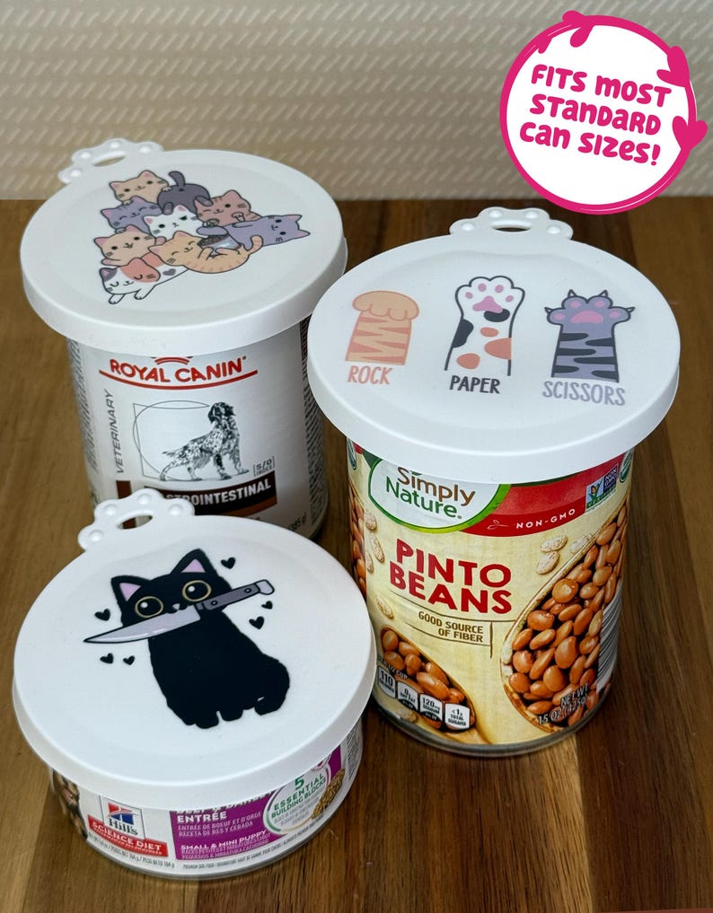 May include: Three white circular silicone can lids with printed designs. One lid features a pile of colorful kittens, another shows cat paws labeled Rock, Paper, Scissors, and the third displays a black cat holding a knife. A pink sticker reads FITS MOST STANDARD CAN SIZES!