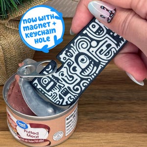 May include: A black and white patterned can opener with a magnet and keychain hole. The opener is being used to open a can of potted meat.