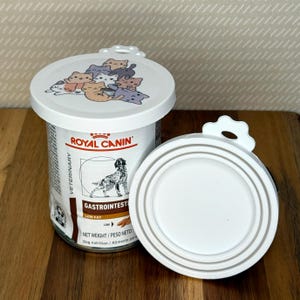 May include: A Royal Canin Veterinary Gastrointestinal Low Fat dog food can with a white plastic lid featuring a cartoon cat illustration, alongside a second identical white plastic lid showing its circular grooved underside design.