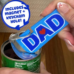 May include: A blue bottle opener with the word "DAD" in white letters and a red heart. The opener is opening a can of food. Text on the image says "INCLUDES MAGNET + KEYCHAIN HOLE!"