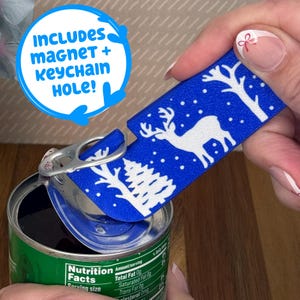 May include: A blue rectangular magnet with a white winter scene of a deer, trees, and falling snow. The text "INCLUDES magnet + keychain HOLE!" is in a blue circle. The magnet is being held over a can.