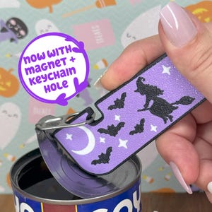 Może przedstawiać: Fioletowy brelok o tematyce Halloween z wiedźmą na miotle, nietoperzami i gwiazdami. Brelok jest trzymany nad puszką. Tekst na breloku brzmi "NOW WITH MAGNET + KEYCHAIN HOLE".