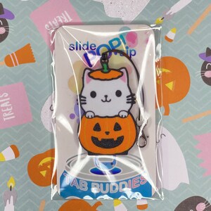 May include: A white cat wearing a pumpkin hat is holding onto a pumpkin with a jack-o'-lantern face. The charm is attached to a black metal slider clip. The charm is packaged on a white card with the text "slide pop! clip" and "Tab Buddies".