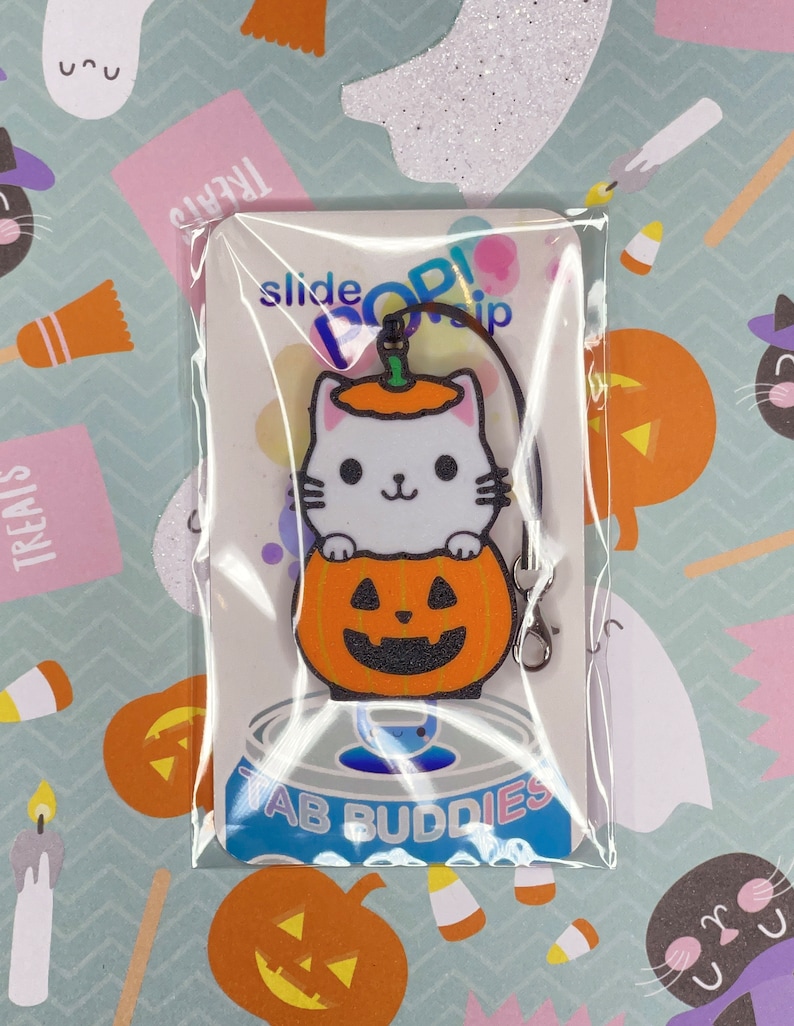May include: A white cat wearing a pumpkin hat is holding onto a pumpkin with a jack-o'-lantern face. The charm is attached to a black metal slider clip. The charm is packaged on a white card with the text "slide pop! clip" and "Tab Buddies".
