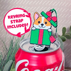 May include: A cute cartoon corgi dog wearing a red and green Santa hat pops out of a green and red gift box. The keychain charm is attached to a silver chain and has a red tag that says "keyring strap included!"