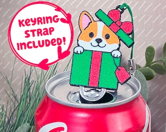 May include: A cute cartoon corgi dog wearing a red and green Santa hat pops out of a green and red gift box. The keychain charm is attached to a silver chain and has a red tag that says "keyring strap included!"