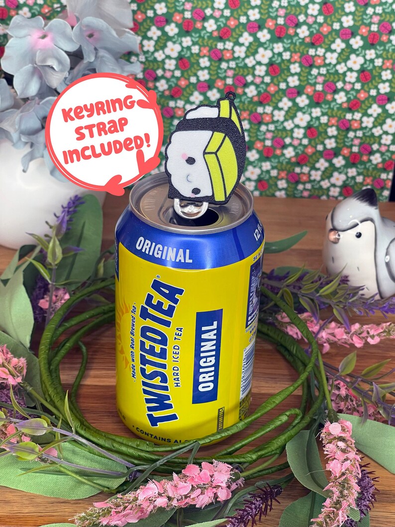 Pu&ograve; includere: Una lattina gialla di Twisted Tea, un t&egrave; freddo forte, con un portachiavi a forma di sushi bianco e nero in cima. Il portachiavi ha un viso da cartone animato carino ed &egrave; attaccato alla linguetta di apertura della lattina. La lattina &egrave; circondata da fiori rosa e viola.