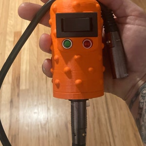 May include: A bright orange, textured, cylindrical handheld device featuring a black rectangular power switch, a small green indicator light, and a small red indicator light. A black cable with a metallic XLR-style connector is attached to the unit.