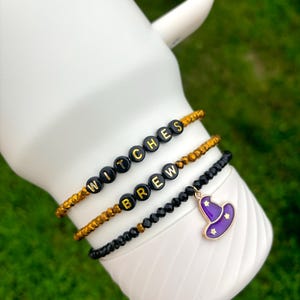 May include: Three beaded bracelets with the words "Witches Brew" spelled out in gold letters on black beads. The bracelets are on a white background. A purple witch hat charm hangs from the bottom bracelet.