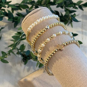 May include: Several beaded bracelets in gold and white are displayed on a beige fabric cylinder. The bracelets feature gold beads, white pearls, and a gold cross charm. Green foliage is visible in the background, adding a natural touch to the presentation.