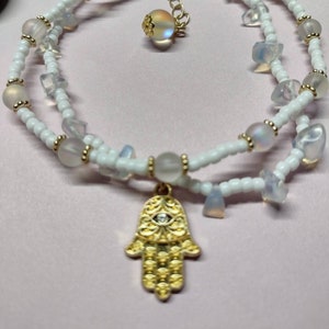 Pode incluir: Pulseira de contas brancas com um amuleto de mão de Hamsa dourado. A mão de Hamsa tem um olho azul no centro. A pulseira é feita com contas brancas pequenas e contas brancas iridescentes maiores.