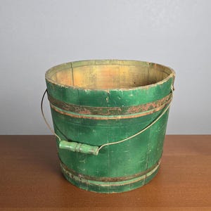 May include: A vintage green wooden bucket with a wire handle and a wooden handle. The bucket has a worn, distressed look and is made of wood slats.