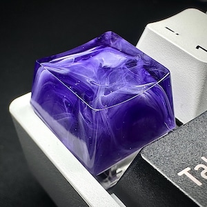 May include: A custom keycap with a swirling purple and white design, set on a mechanical keyboard. The keycap is square and translucent, with a marbled effect. The keyboard keys are white and black.