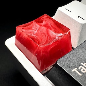 May include: A custom red and white keycap with a marbled design, installed on a mechanical keyboard. The keycap is square and sits between a white key with the number 1 and a black key labeled "Tab".