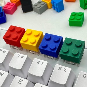 May include: A selection of colourful brick-shaped keycaps in red, yellow, blue, and green, placed on a white keyboard. Other keycaps in various colours are visible in the background. The keycaps are designed to look like toy building blocks.