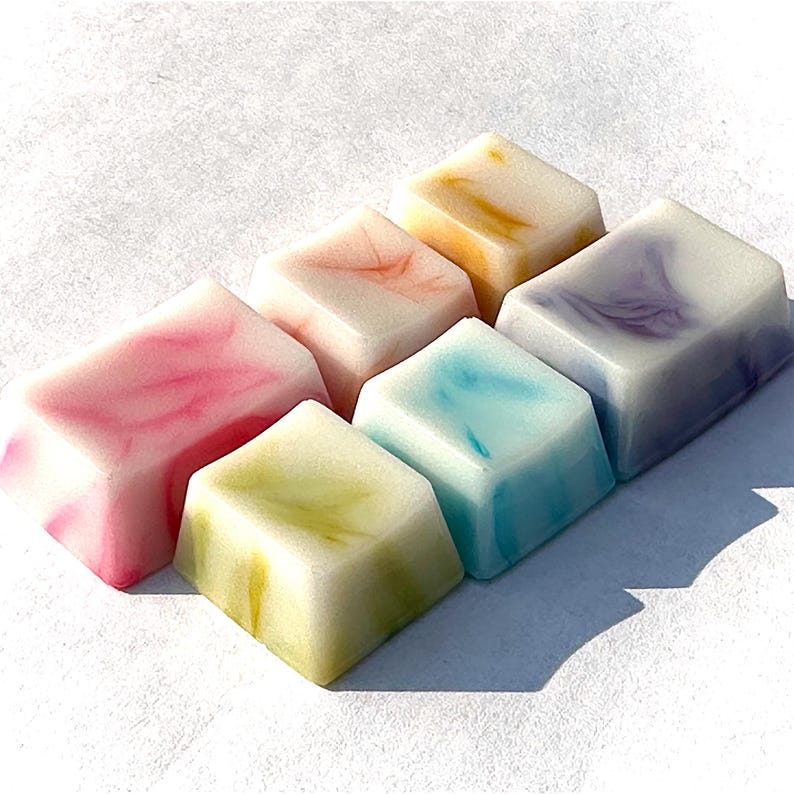 Pu&ograve; includere: Sei saponette quadrate e marmorizzate di colori vivaci. Le saponette sono disposte in fila, con la saponetta pi&ugrave; grande a sinistra e la pi&ugrave; piccola a destra. I colori delle saponette sono rosa, giallo, arancione, blu, viola e verde.