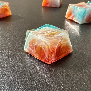 Pu&ograve; includere: Un set di cinque tasti in resina colorati con un motivo a spirale di bianco, arancione e blu. I tasti sono quadrati e hanno una finitura lucida.