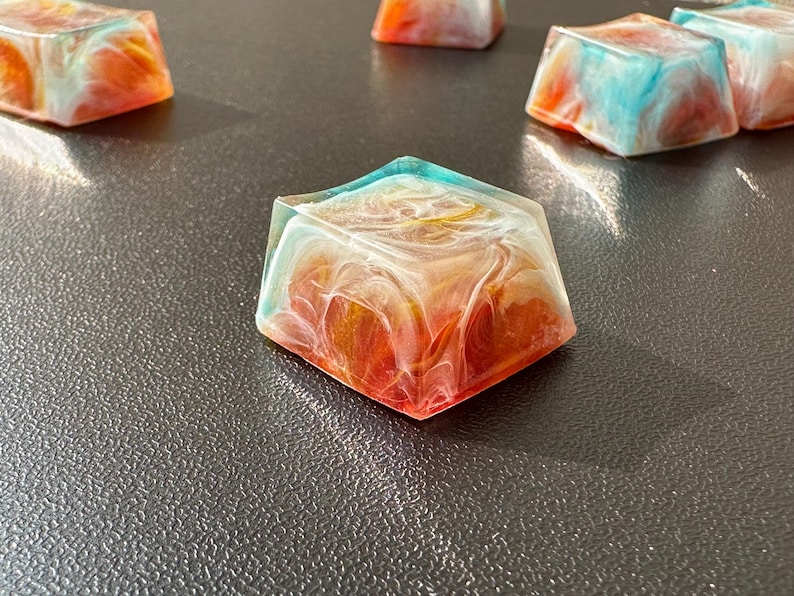 Pu&ograve; includere: Un set di cinque tasti in resina colorati con un motivo a spirale di bianco, arancione e blu. I tasti sono quadrati e hanno una finitura lucida.