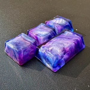 Pu&ograve; includere: Quattro tasti quadrati in resina con un design a spirale galattica in tonalit&agrave; di viola, blu e bianco.