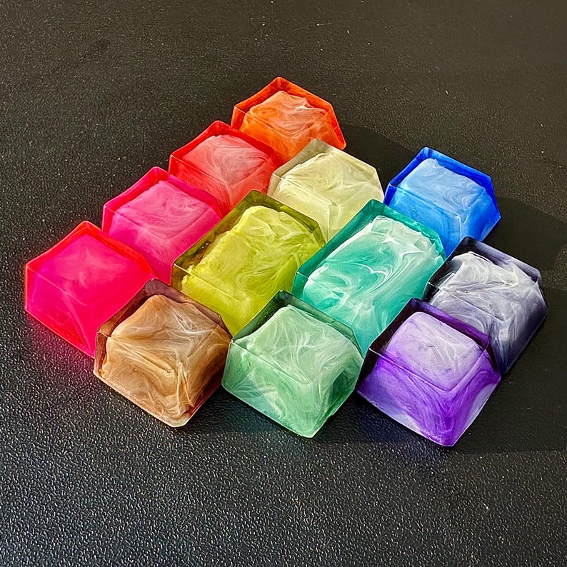 Pu&ograve; includere: Un set di 10 tasti in resina colorati con un motivo a spirale. I tasti sono disposti a forma di triangolo e sono realizzati in un materiale traslucido. I colori includono rosa, verde, blu, viola, marrone, giallo e bianco.