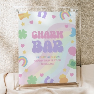 May include: A clear acrylic sign with a colorful design that reads "Charm Bar". The sign features pastel rainbows, stars, hearts, and shamrocks. The text below says "Create your own charm bracelet or necklace".