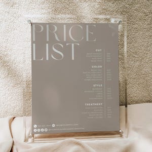 Peut inclure: Un porte-affiche en acrylique transparent contenant une liste de prix grise pour des services de salon. Le texte inclut des catégories pour la coupe, la couleur, le style et le traitement, avec des coordonnées en bas. La marque BGBLOSSOM est en haut.