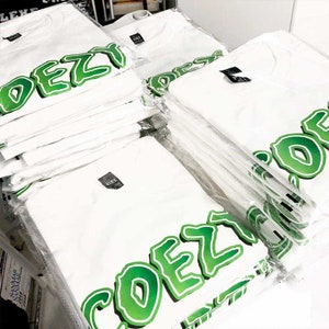 May include: A stack of white t-shirts, each packaged in clear plastic. The shirts feature the word "DEZY" in large, green, bubble-style letters. Each shirt has a black tag.