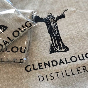 May include: Tan linen fabric with the words "Glendalough Distillery" printed in black. A black graphic of a figure with outstretched arms is also printed on the fabric. Two packaged items are visible in the image.