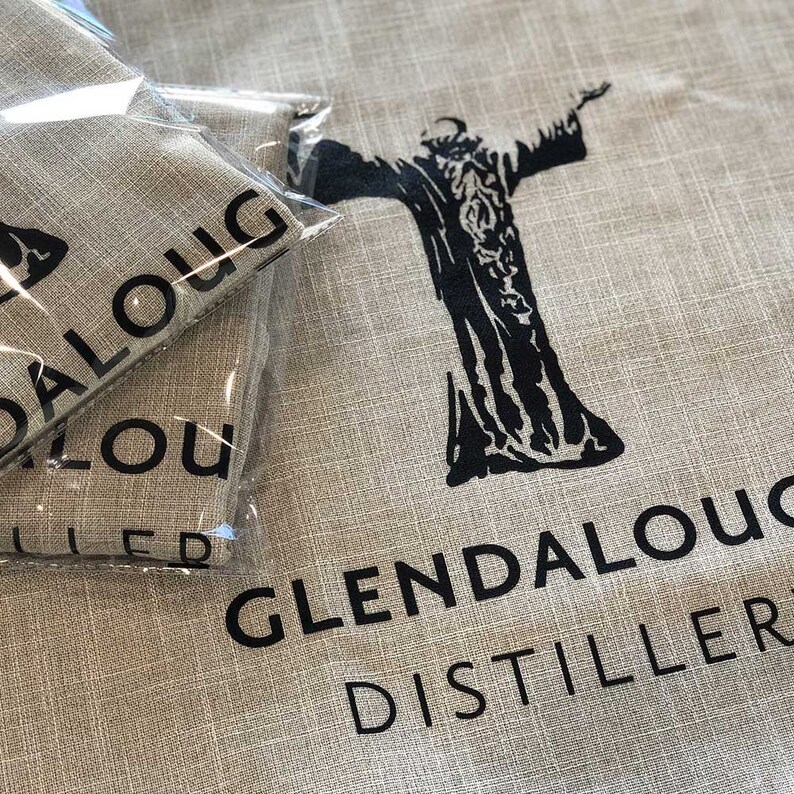May include: Tan linen fabric with the words "Glendalough Distillery" printed in black. A black graphic of a figure with outstretched arms is also printed on the fabric. Two packaged items are visible in the image.