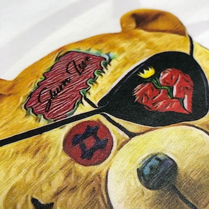 May include: A yellow teddy bear with a black eye patch and a red heart with a crack in it. The bear has a red tattoo that says "Gummy Love" and a red tattoo with a blue and black symbol.