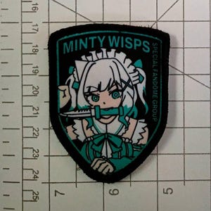 Puede incluir: Parche negro y verde azulado con una ilustración de personaje de estilo anime. El personaje tiene cabello blanco y ojos verde azulado, sosteniendo un cuchillo. El texto en el parche dice "MINTY WISPS" y "SPECIAL FANDOME GROUP".