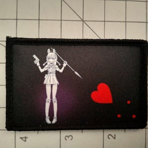 May include: Black and white embroidered patch featuring a cartoon character holding a gun and a spear. The character is wearing a skirt and boots. The patch also includes a red heart and three red dots.