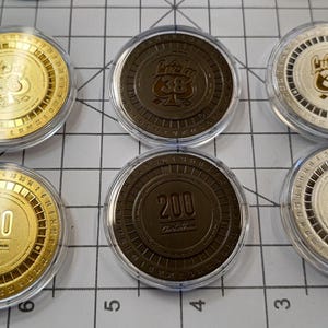 Könnte beinhalten: Sechs runde Casino-Chips in durchsichtigen Plastikbehältern. Die Chips sind golden, silbern und schwarz mit verschiedenen Designs. Ein Chip hat die Nummer 200 mit dem Text "On the House". Ein weiterer Chip hat die Nummer 200. Ein weiterer Chip hat den Text "Lucky 38" darauf. Die Chips befinden sich auf einer gerasterten Oberfläche.