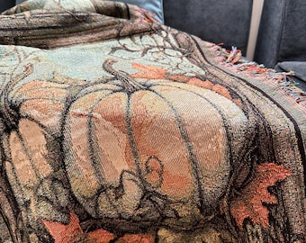 May include: A woven throw blanket featuring a detailed autumnal scene with large orange pumpkins and fallen maple leaves in shades of red, orange, and brown. The textile has a decorative border and a fringed edge with multi-colored threads.