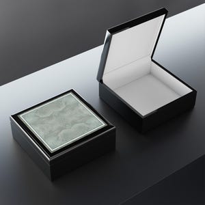 May include: Two square black boxes, one closed and one open. The closed box features a textured, light gray square panel on its lid. The open box reveals a plain white interior. Both boxes have a smooth, dark finish and a clean, geometric design.