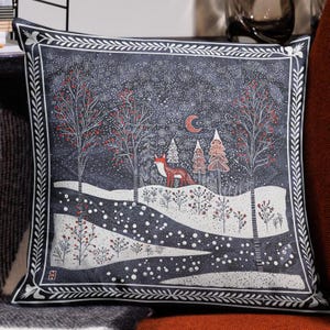 May include: A dark blue throw pillow with a white border features a winter scene with a red fox walking through a snowy forest. The forest is filled with white trees and red berries. The background is a dark blue sky with white snowflakes.