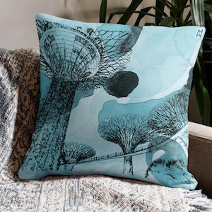 May include: Square throw pillow with a teal and black illustration of stylized trees and a bridge. The pillow has a watercolor effect with a light blue background. The pillow is on a neutral-colored couch.