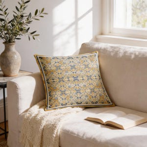 May include: Square decorative pillow with a yellow and blue floral pattern, placed on a cream-colored sofa. The pillow has a dark blue border and a yellow trim. A beige throw blanket and an open book are also on the sofa.