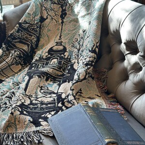 Könnte beinhalten: Eine gewebte Wohndecke mit einem Landschaftsmotiv in Schwarz, Creme und Blaugrün, das eine zentrale Pagode und Bäume zeigt, kombiniert mit einem aufgeschlagenen, dunkelblauen Hardcover-Buch mit dem Titel Poems und goldener Schrift.
